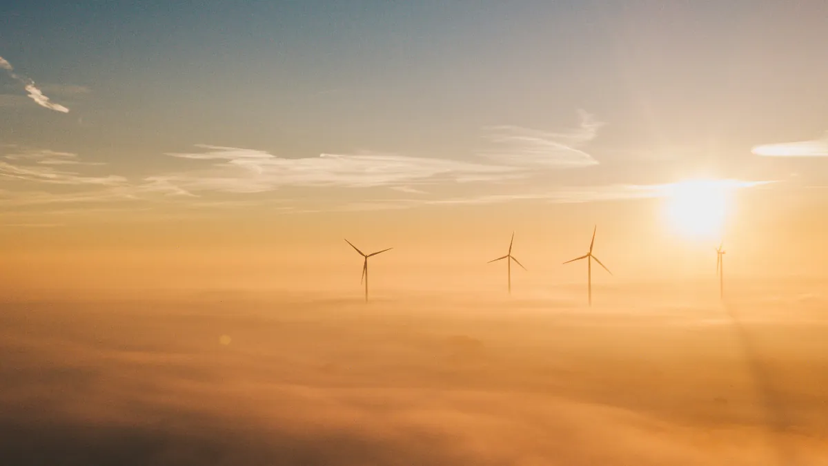 three electricity-generating windmills poking through the top of clouds, with the sun shining on the right, giving the clouds an orange hue